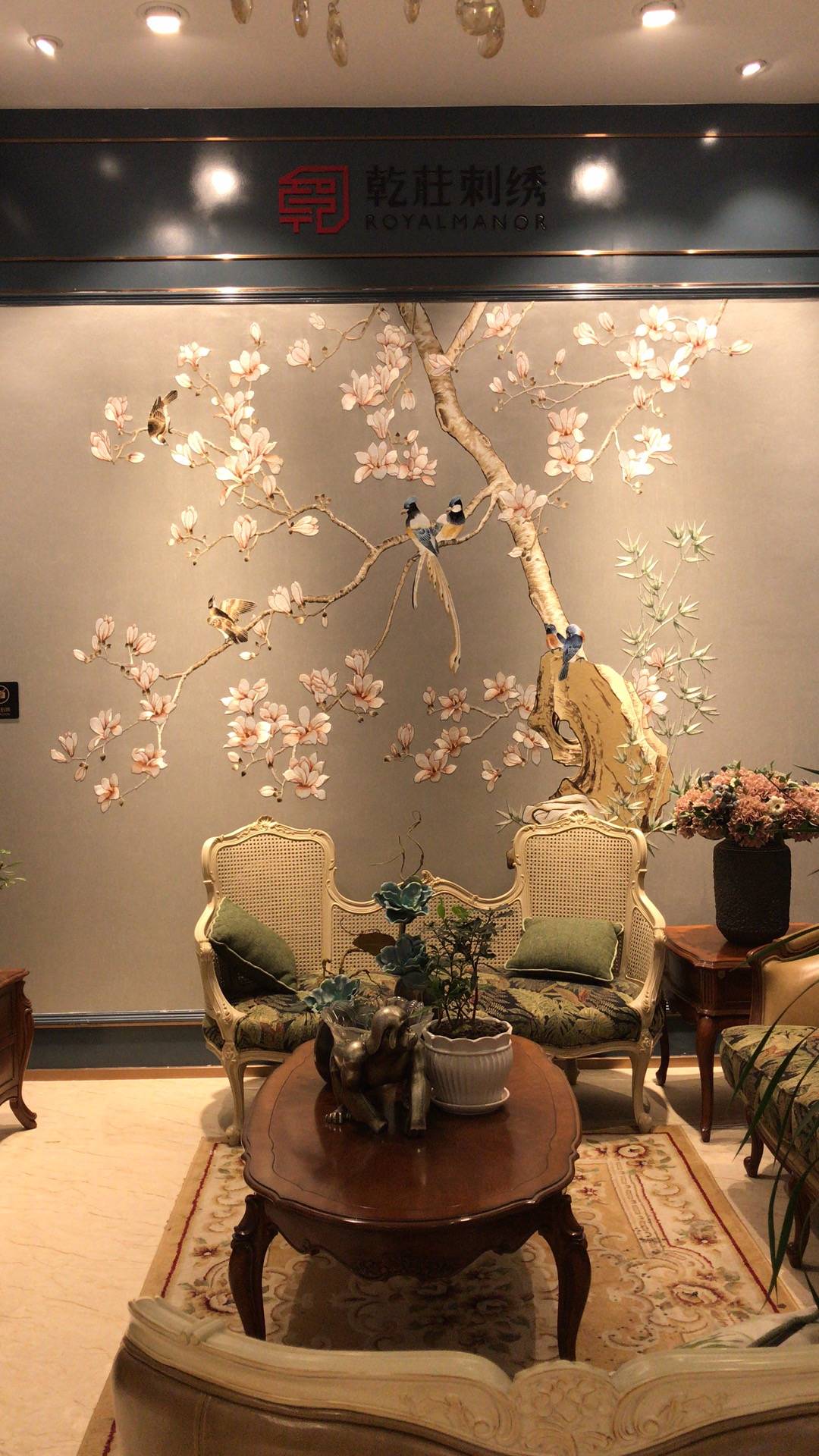 乾莊刺繡展厅实景，背景墙展示大幅玉兰花鸟刺绣壁画，工艺精湛，色彩柔和。搭配法式藤编沙发与深色实木茶几，绿色靠枕点缀，营造出新中式与欧式融合的典雅氛围。适合高端别
